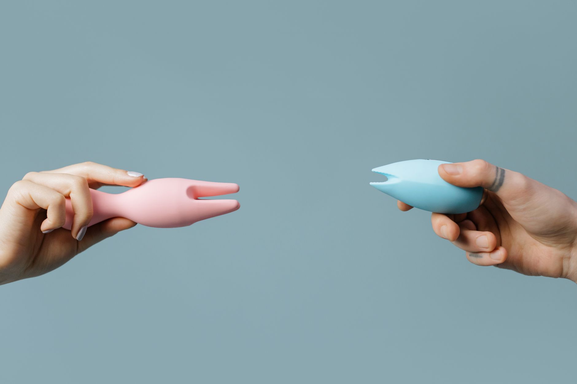 Two hands holding colorful silicone vibrators against a pastel background